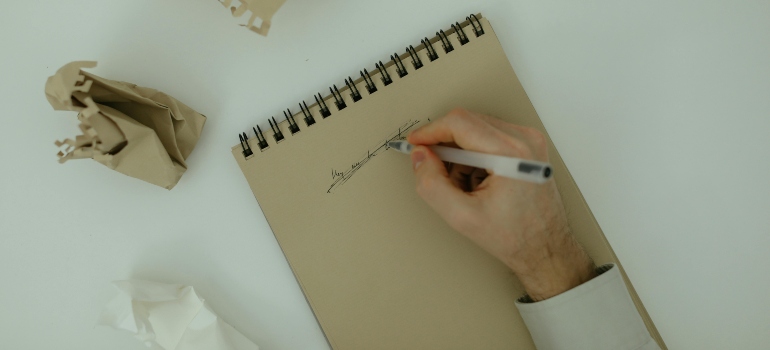 Person crossing out notes in a spiral notebook surrounded by crumpled paper.