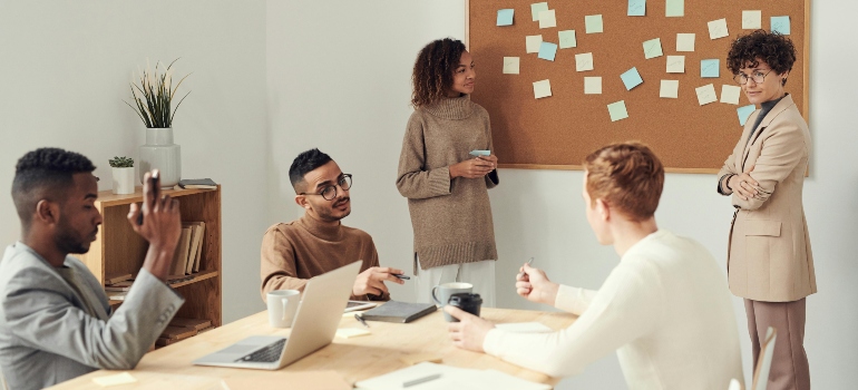 Team gathered around a table in discussion with sticky notes on the wall in a bright office.
