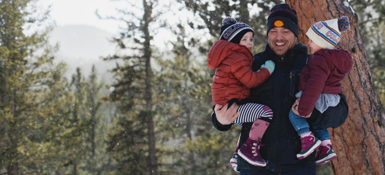 Father holding two kids in a snowy Colorado forest, everyone bundled up in colorful winter hats.