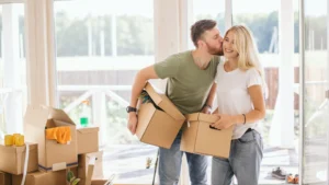 A happy young couple kiss while carrying brown cardboard moving boxes as part of their DIY move.