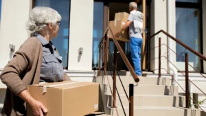 An elderly man and woman carry cardboard boxes of their belongings.