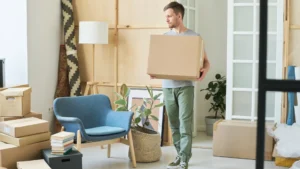 A man carries a brown paper box to the other side of the room while waiting for long-distance movers to arrive.